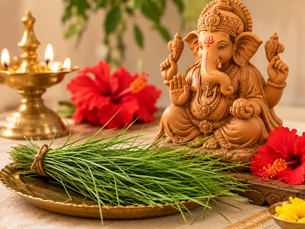 Fresh green durva grass arranged as a traditional offering beside a clay Lord Ganesha idol, red hibiscus flowers and a brass diya.